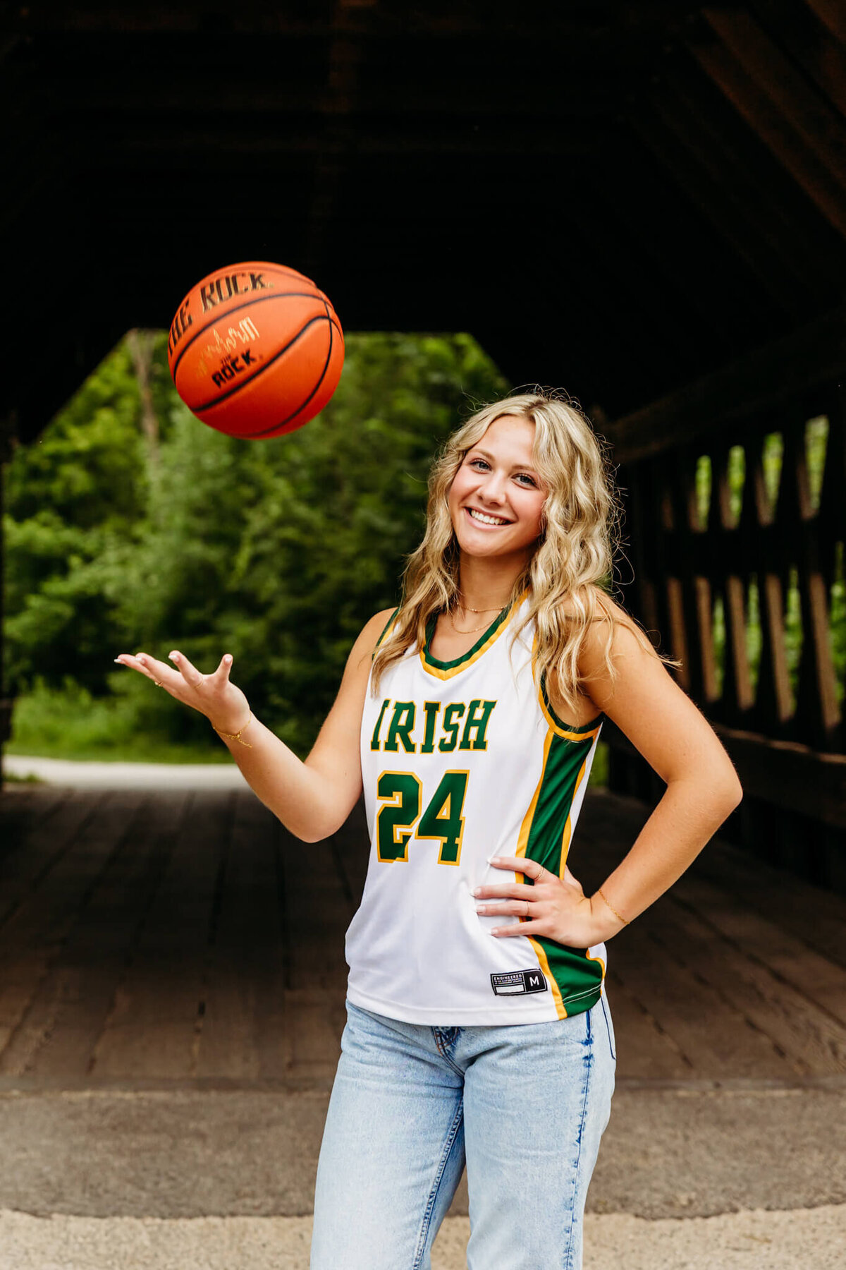 blonde teen girl in a basketball jersey tossing a basketball up in the air and smiling for her senior photos.