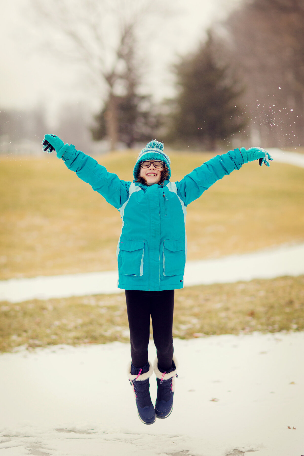 Girl is bundled up in blue coat and winter boots, jumping in the snow.