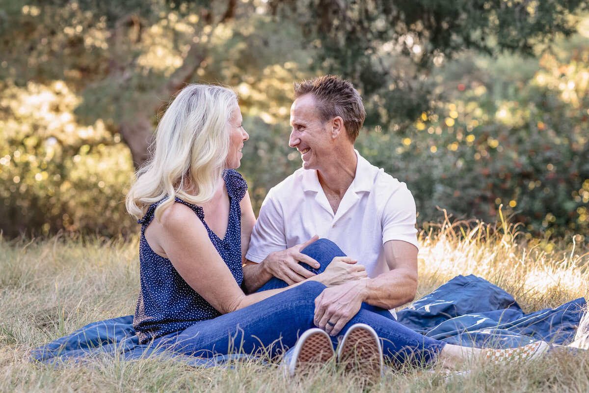 A husband and wife sitting on a picnic blanket in the grass, looking at each other and smiling, surrounded by trees.