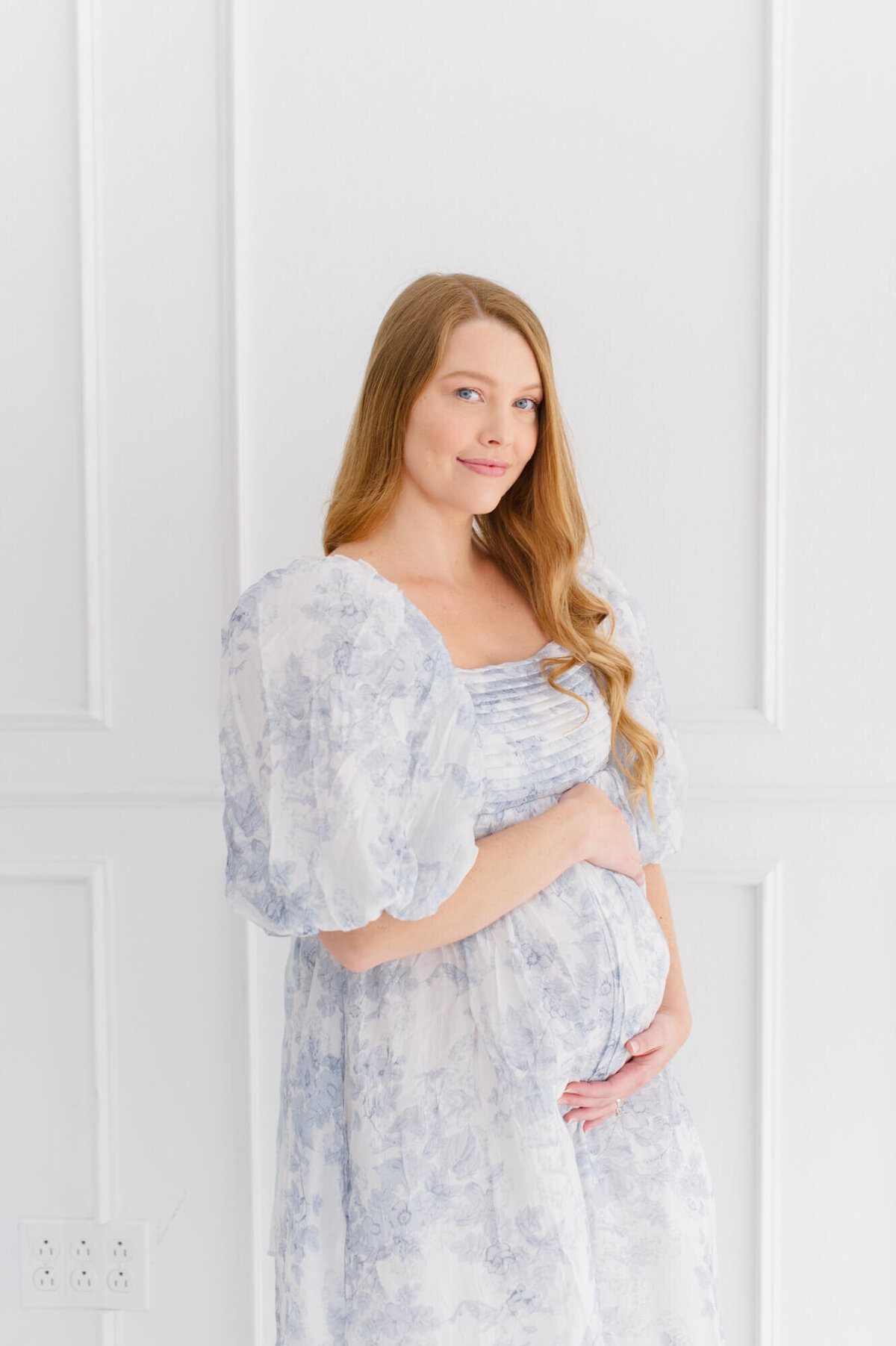 Pregnant mom standing on white wall posing and smiling at the camera