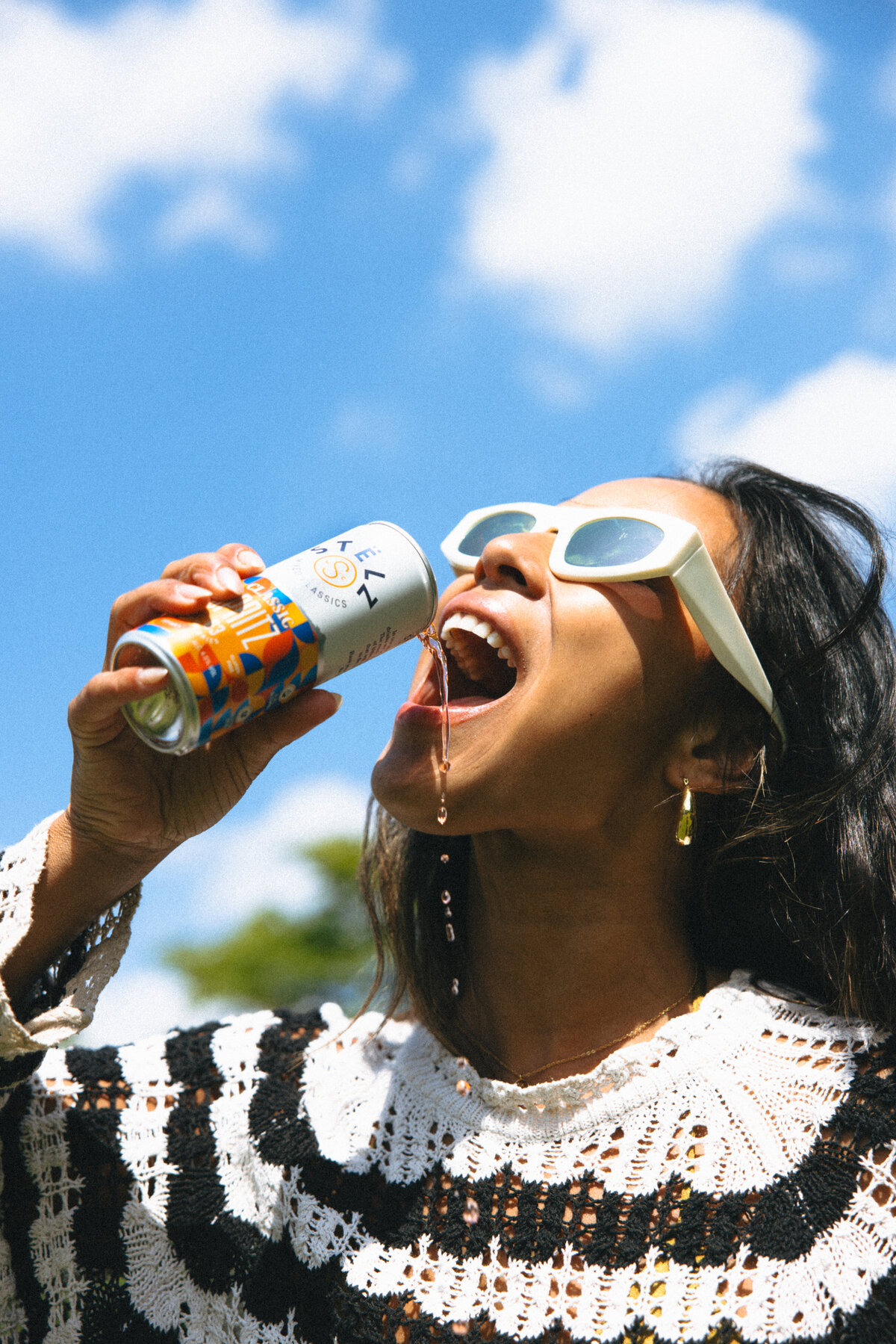 A woman enjoys her STËLZ Classic Spritz, holding the can high above her head as she takes a sip. The blue sky and her joyful expression amplify the summer and festive feeling of the photo. Photo taken by Bo Eppink.