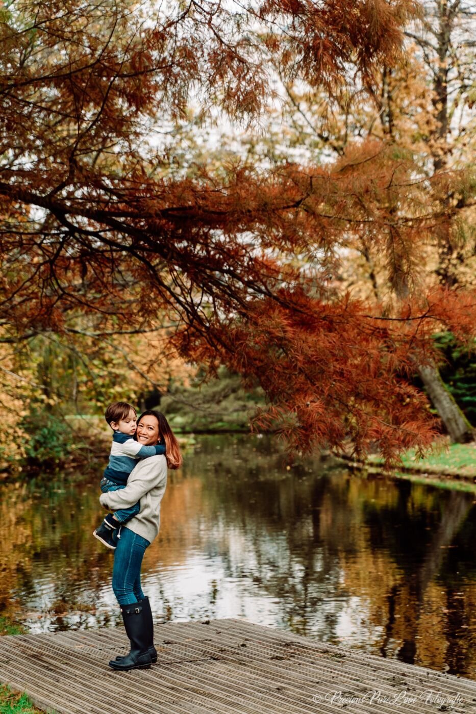 Mother holding son in autumn park – Woman wearing boots and sweater carrying her young son on a wooden platform by a pond framed with golden fall foliage.