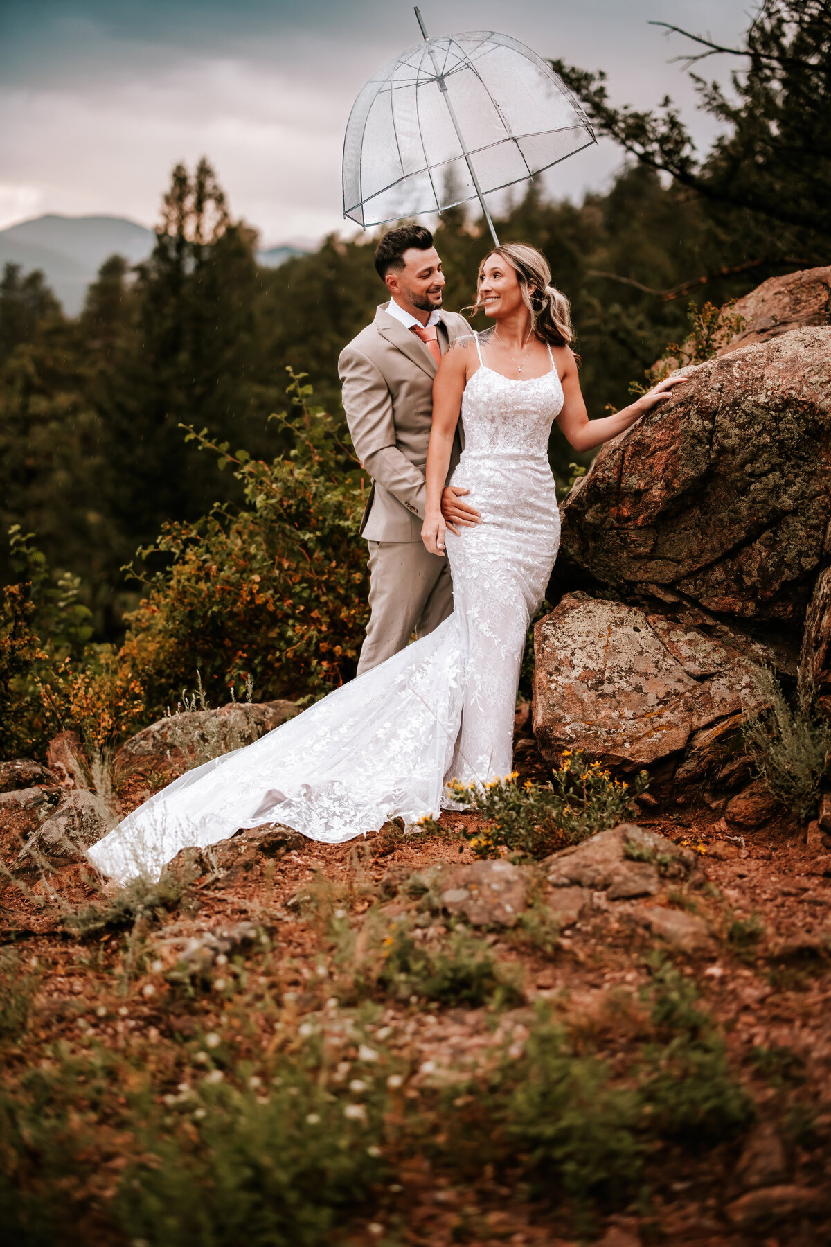 Husband embracing wife from behind while leaning against a rock and holding a clear umbrella