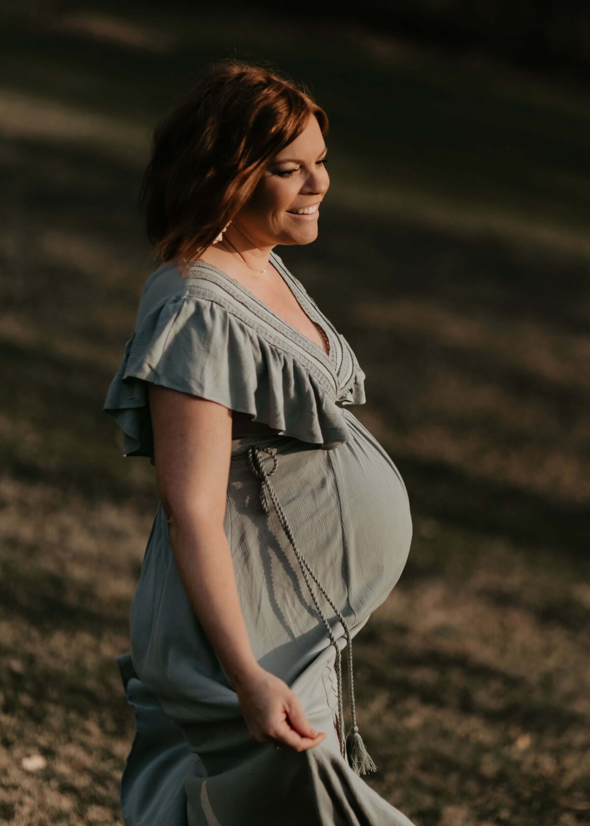 A pregnant woman in a blue dress is strolling gracefully through a field, captured by a Pittsburgh maternity photographer.