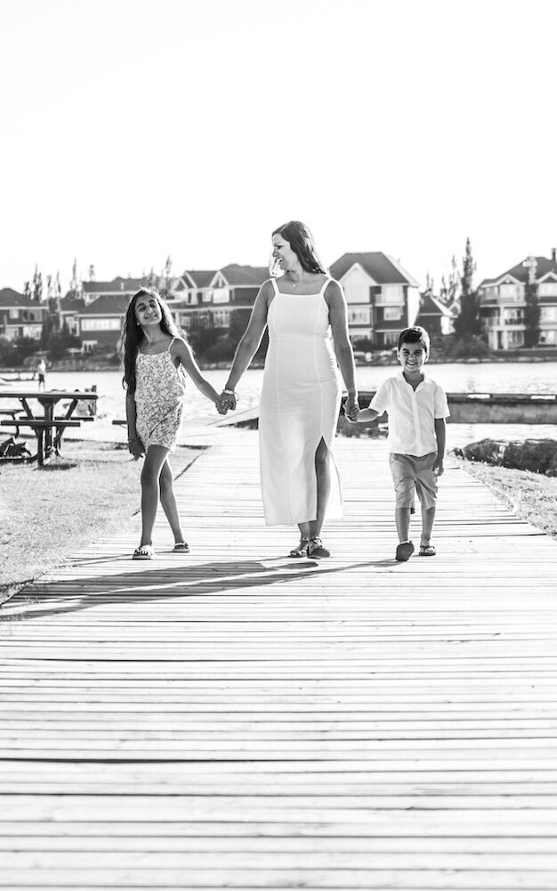 A mom in a white dress is walking near a lake holding her son and daughter's hands