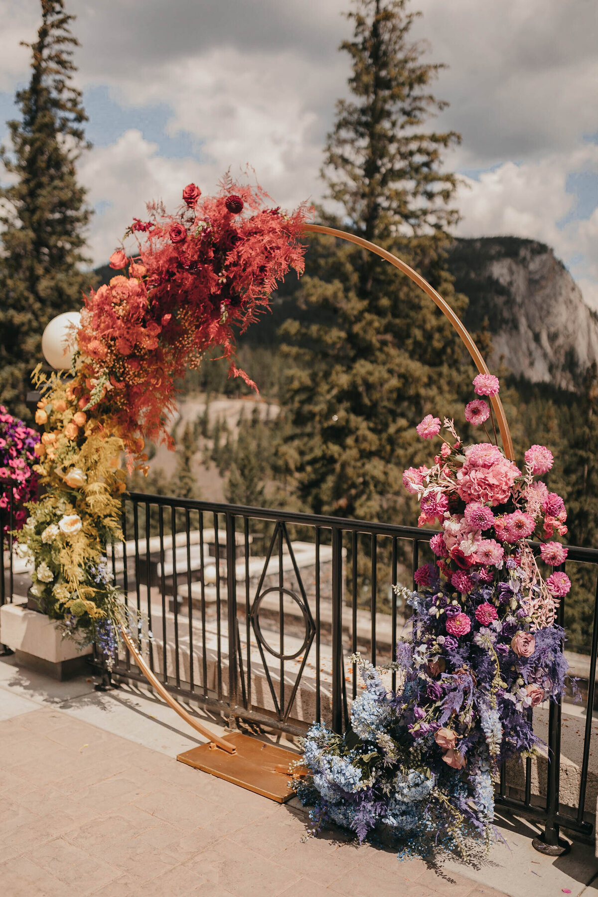 A semi-circular metal arch adorned with colorful flowers stands on a terrace overlooking a forested mountain landscape, setting the perfect scene for a modern day fairytale wedding at Banff Springs Hotel in Banff Canada.