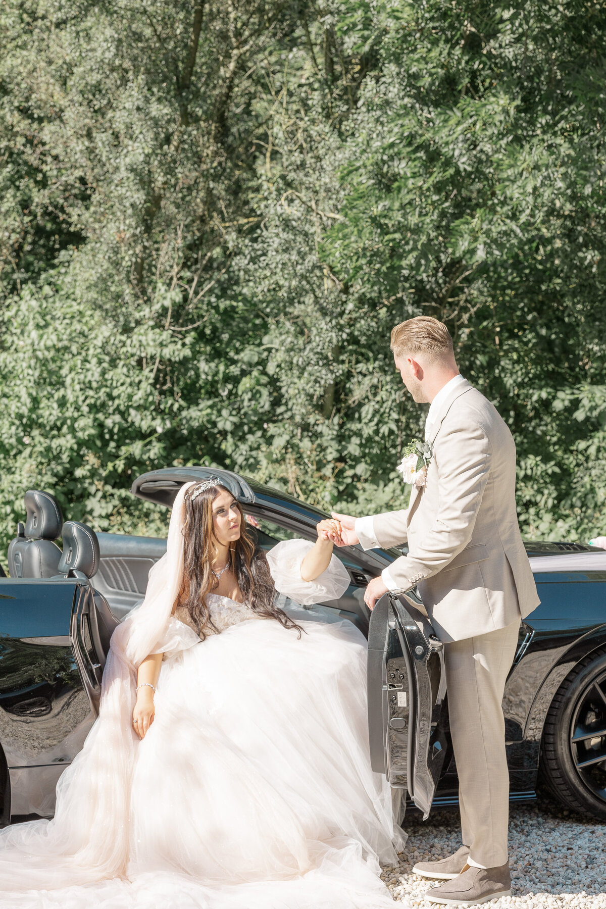 A bride in a voluminous designer dress is being helped out of a convertible mustang by her groom  