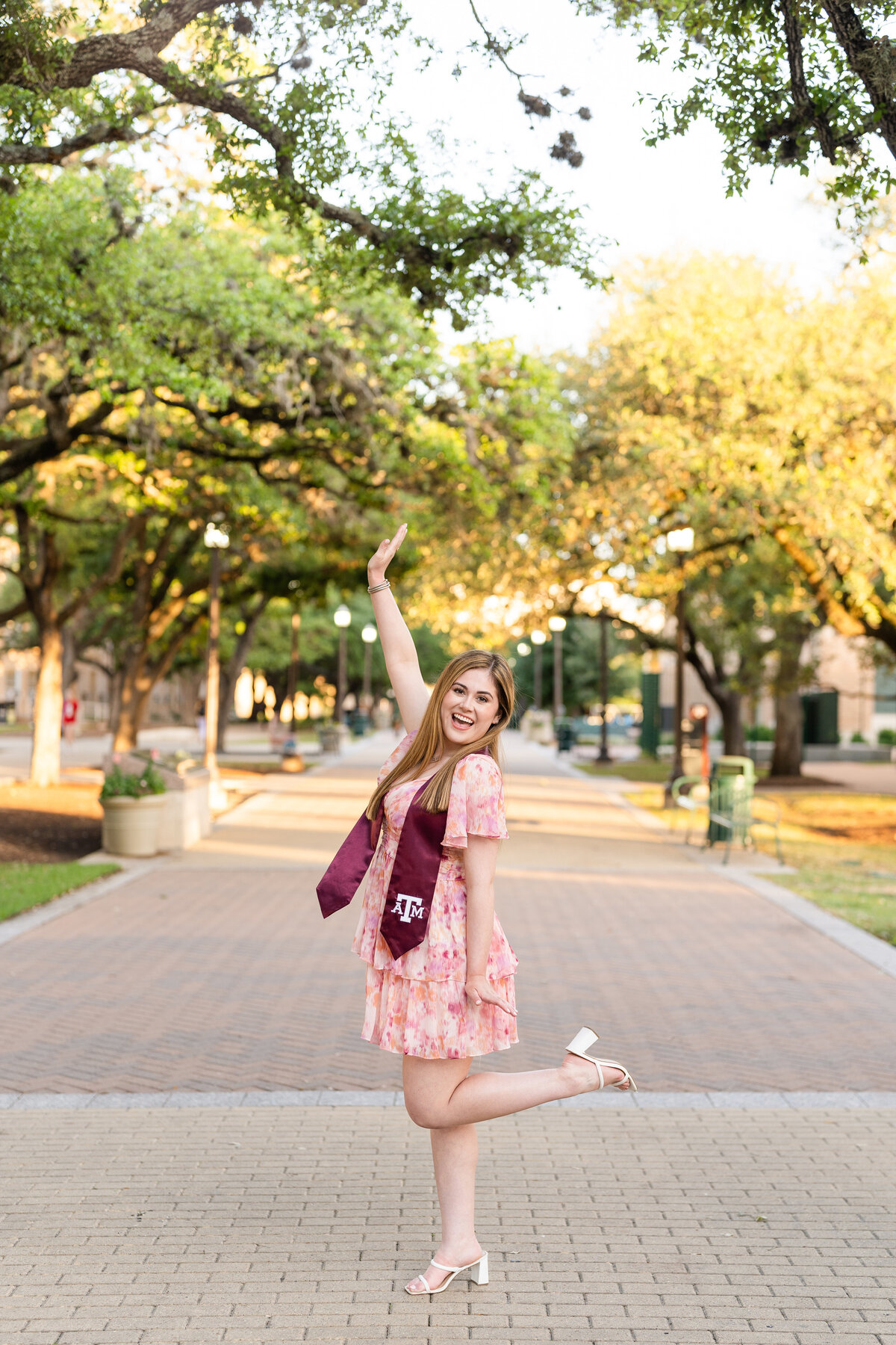 Texas A&M senior girl celebrating with hands in the air while wearing Aggie stole and multi colored dress in the trees of Military Plaza