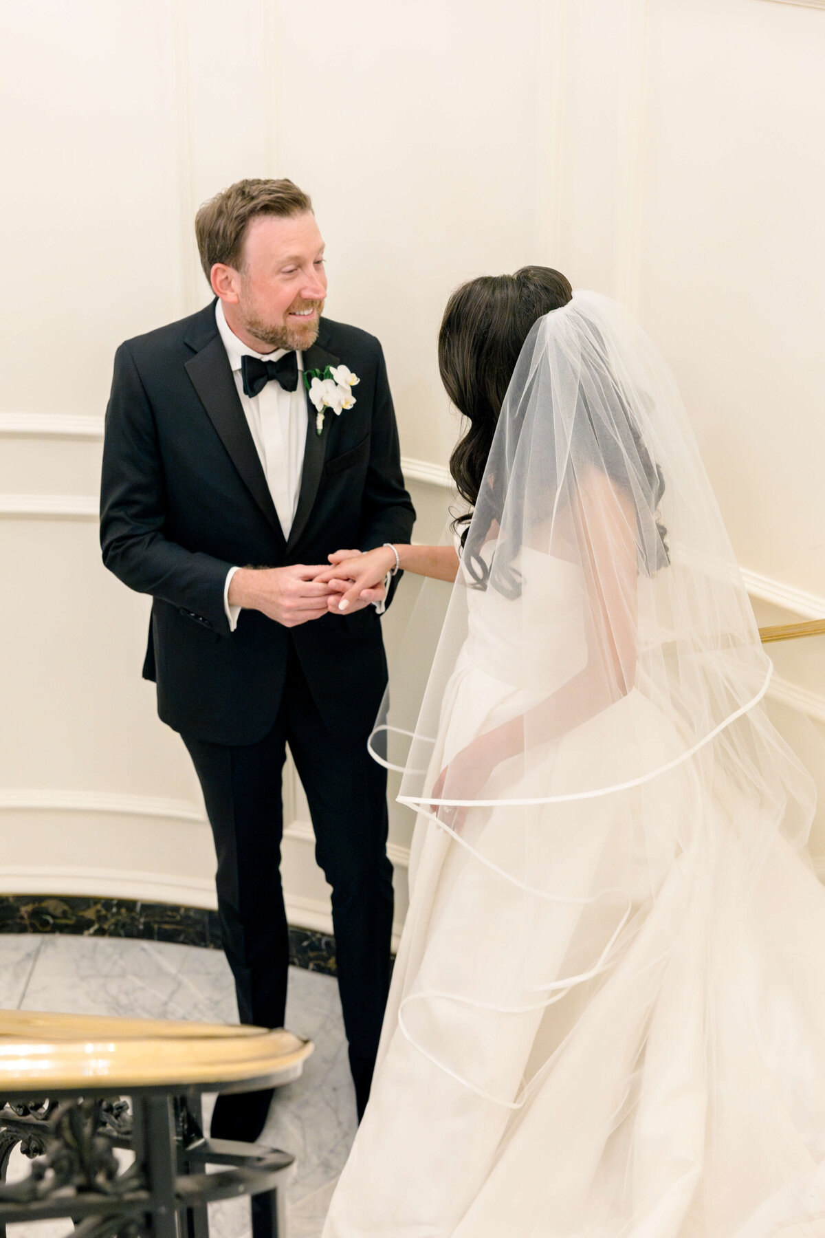 A groom in a black tuxedo and bow tie holds hands with a bride in a white gown and veil, standing together on a staircase with white walls. Both appear joyful and are looking at each other.