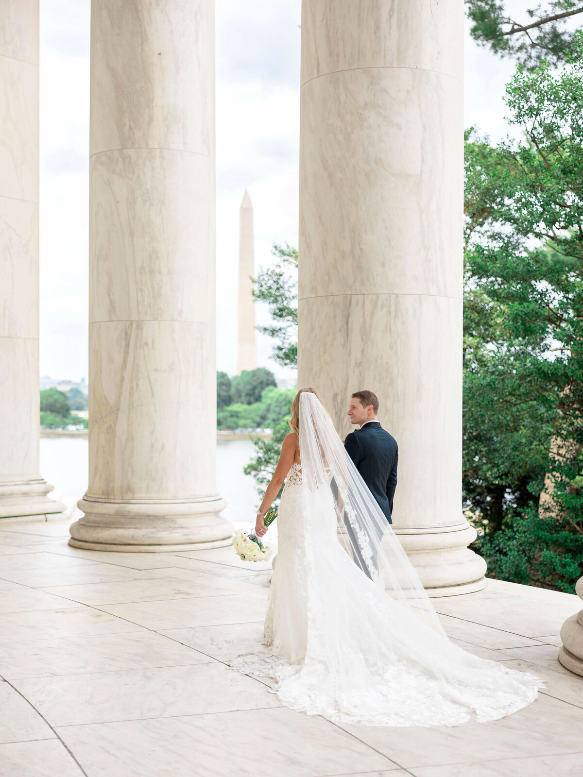 A bride and groom stand together under large marble columns, overlooking a scenic view with the Washington Monument in the background. The bride is in a white dress with a long veil, holding a bouquet.