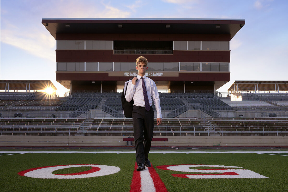A senior in a suit walks confidently on the football field at Mustang-Panther Stadium, captured by a Dallas senior photographer during golden hour.