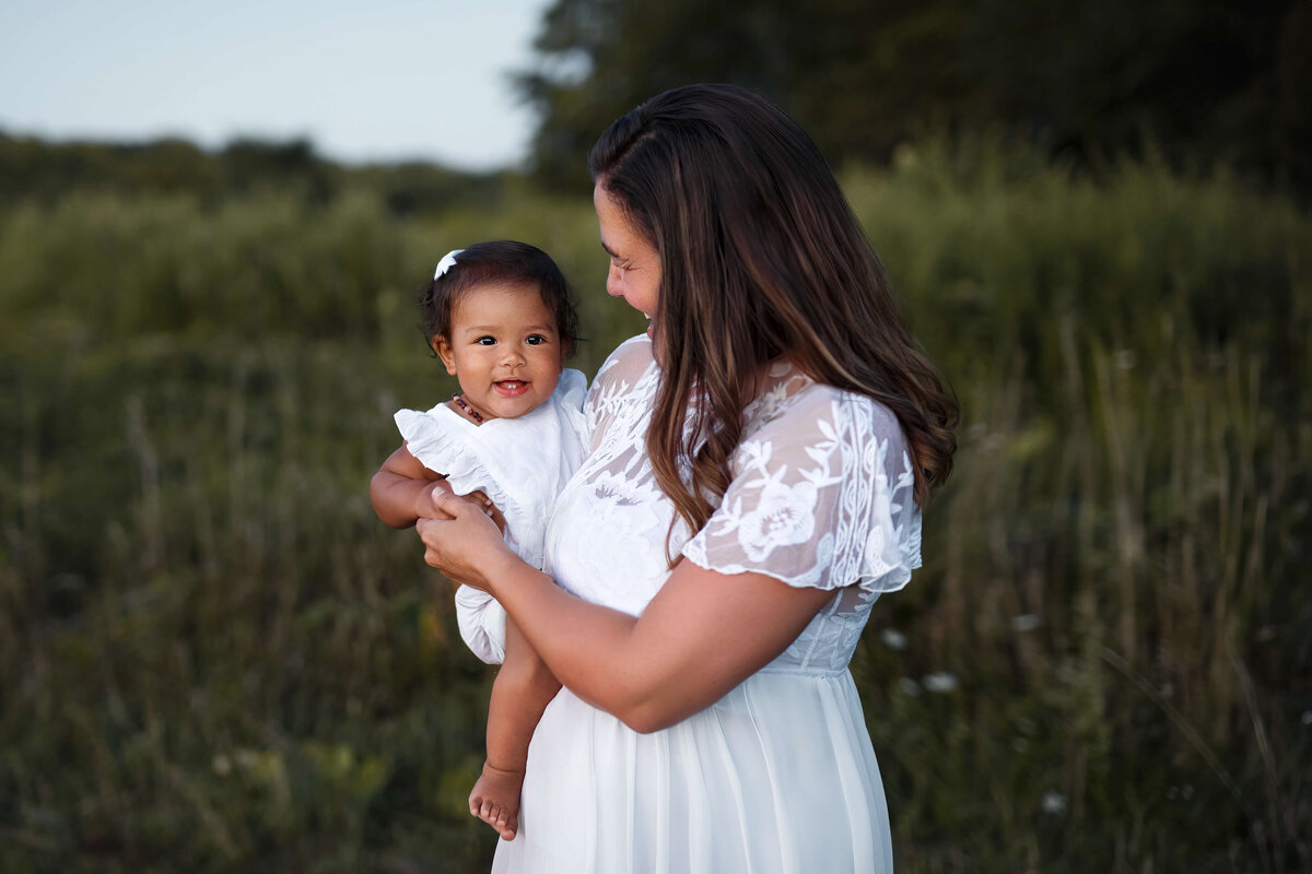 young mom looking at her little girl wearing white dresses in front of a grassy field at sunset in the summer