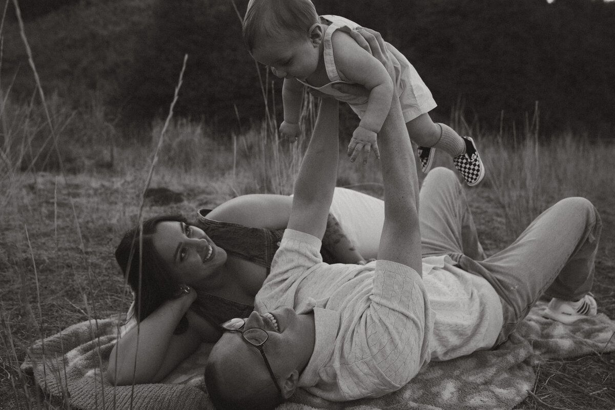 A dad is holding the hand of his toddler and the edge of a lake. The wife is behind, holding their baby.