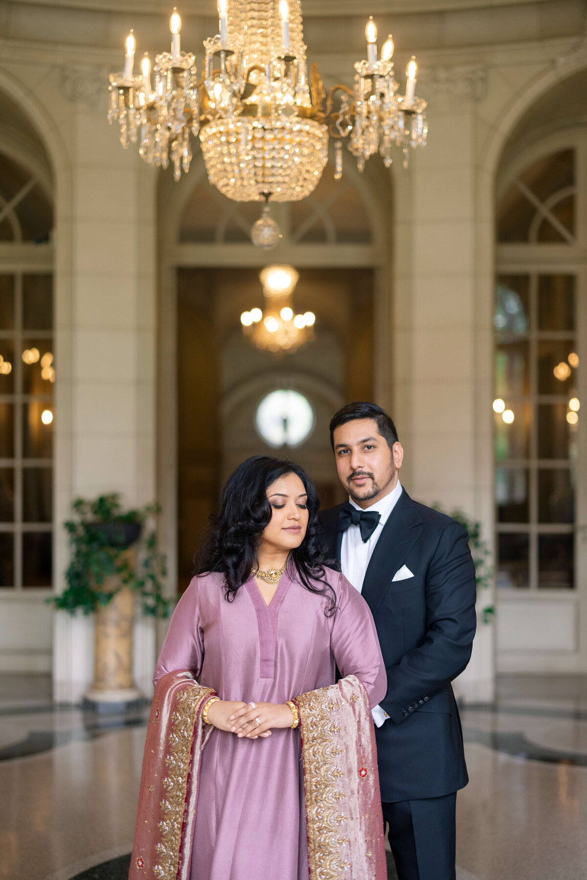 A man in a tuxedo and a woman in a purple gown with a matching shawl stand together in an elegant room with a large chandelier and arched windows.