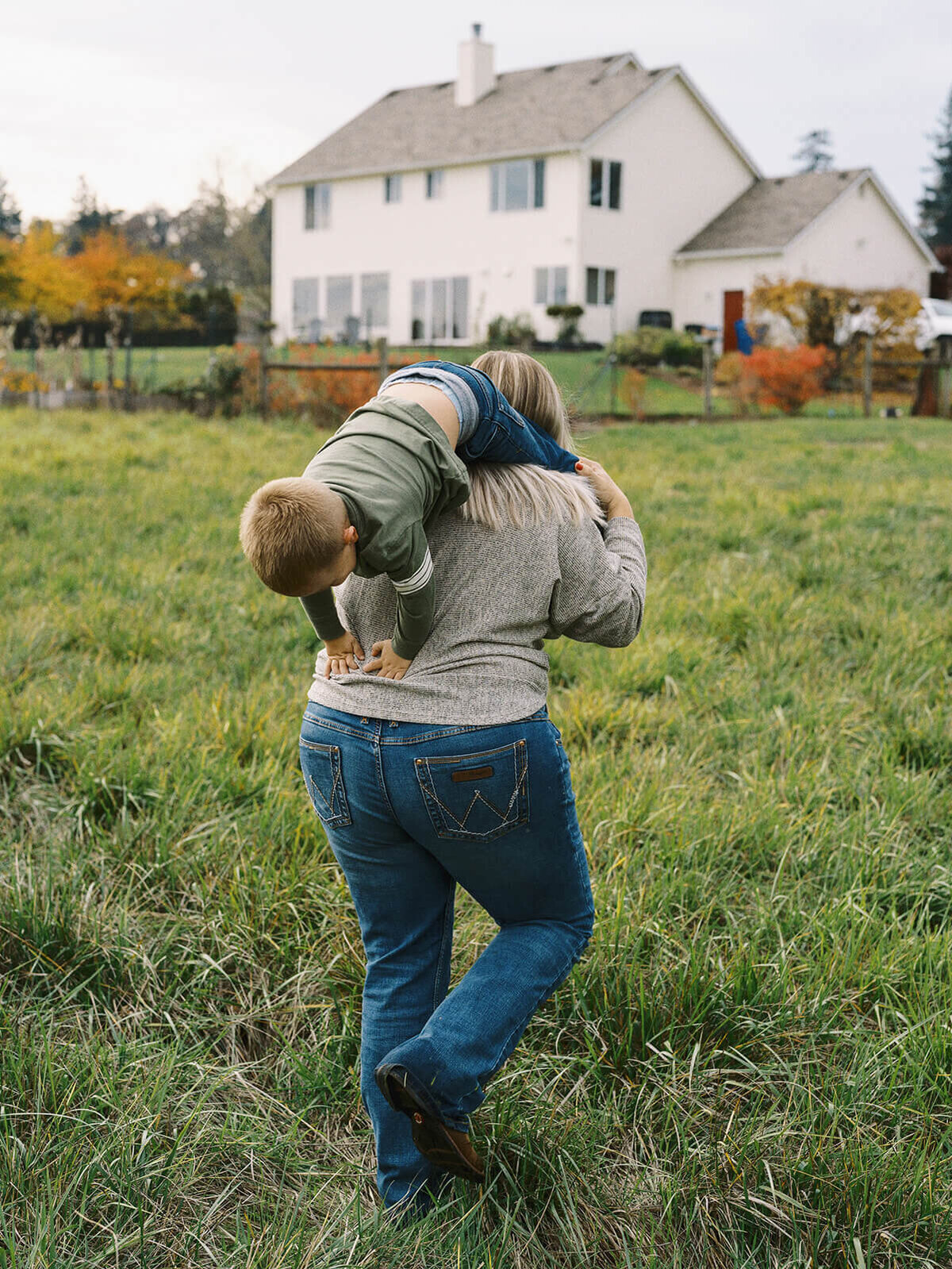 A mom carrying her son over her shoulder while walking away from the camera with their home in the background.