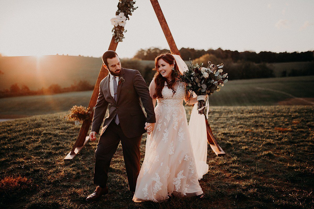 Groom wearing a dark maroon suit and a bride in a long lace dress and veil holding a large bouquet of greenery and dar red flowers stand in front of a triangle arbor decorated with white flowers and the sun sets in the Tennessee mountains