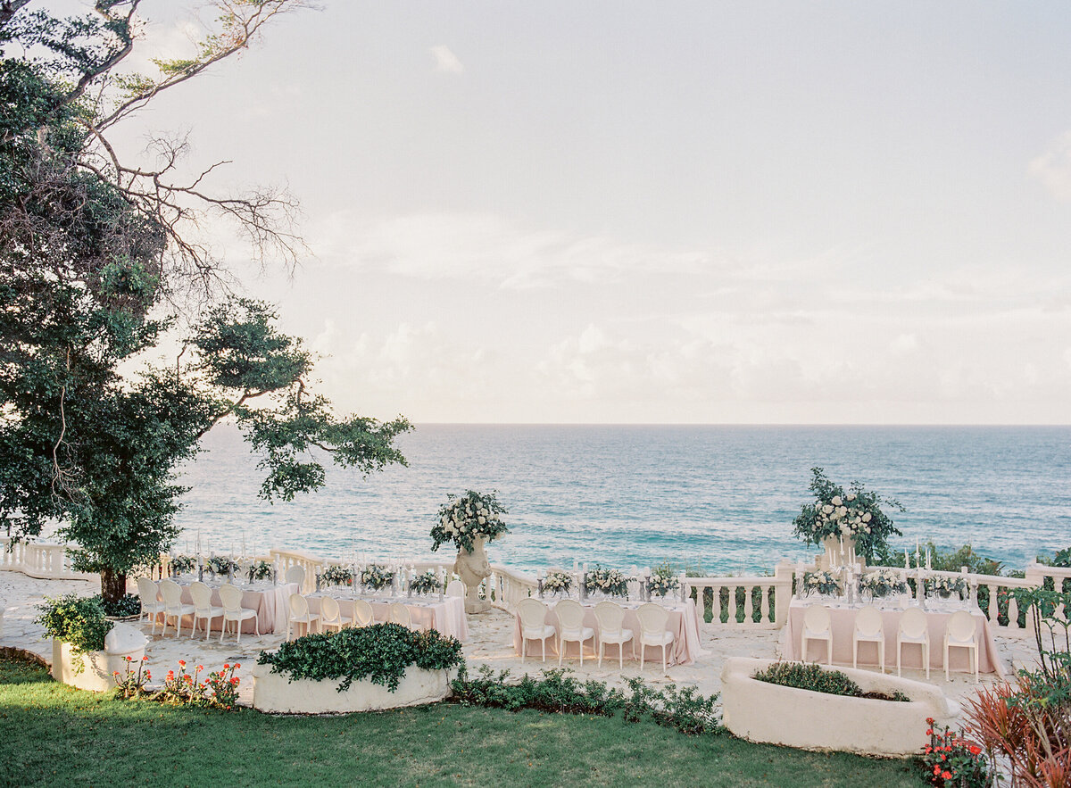 Outdoor wedding reception setup at a top tropical destination wedding venue in the Caribbean, Dominican Republic. White chairs and tables arranged in a U-shape on a terrace with floral centerpieces, overlooking the ocean. Captured by photographer Laura Gordon, creating a serene and elegant ambiance.