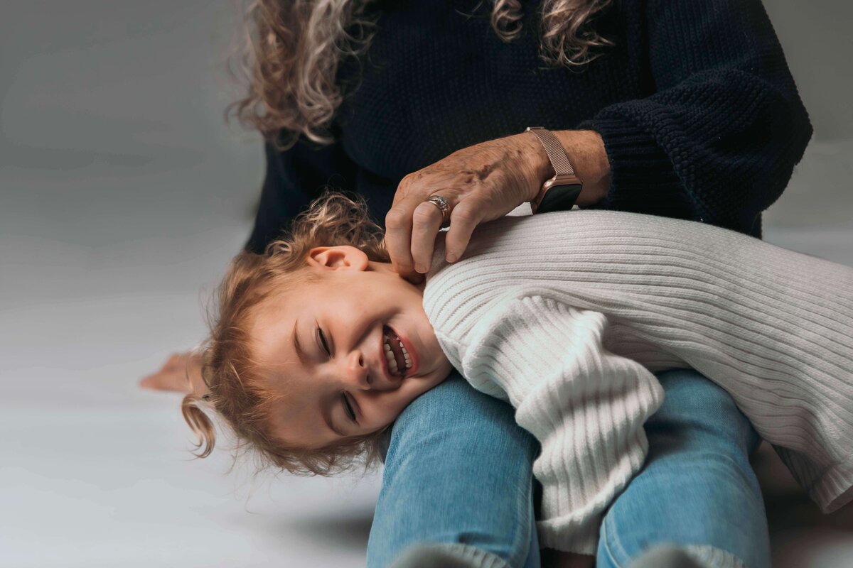 A grandmother in a cozy outfit of blue jeans, a black sweater, and grey socks sits on the floor of Mobile, AL Motherhood Photographer’s studio, tickles  her 2-year-old granddaughter, who is wearing an oversized white sweater onesie, as she lays across her grandmother's legs. The playful portrait captures their personality and bond.