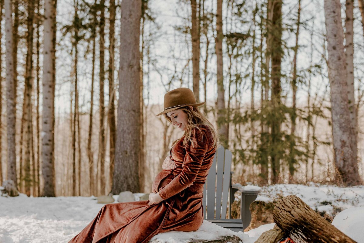 Outdoor winter maternity photoshoot in London, Ontario - Expectant mom sitting side profile to the camera on a bench. She is wearing a long burnt orange gown, holding and looking down at her baby bump. Tall trees are in the background and the grown is snow covered.