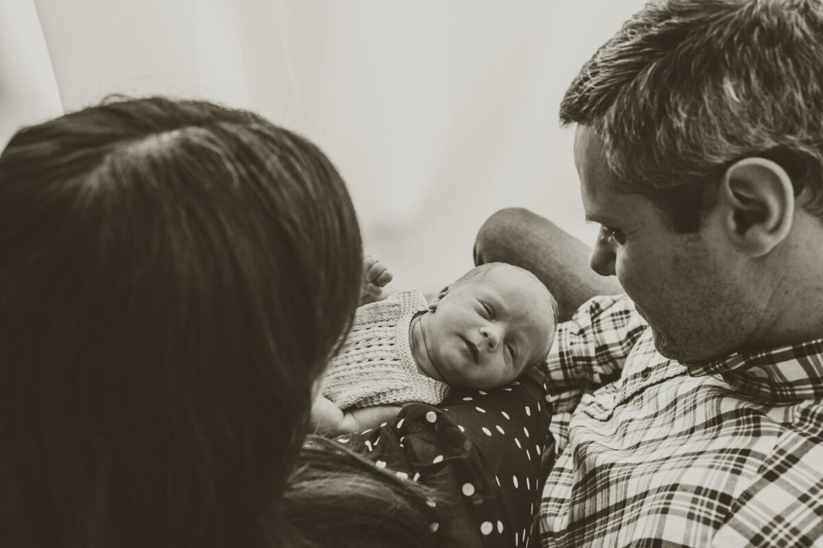 A tender monochrome moment capturing a father and mother looking with love at their peacefully sleeping newborn baby nestled between them