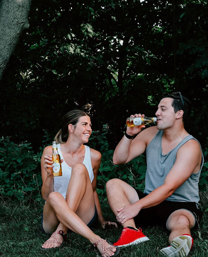 A man and woman chill on the grass, each holding a Michaelob Ultra beer, smiling and chatting.