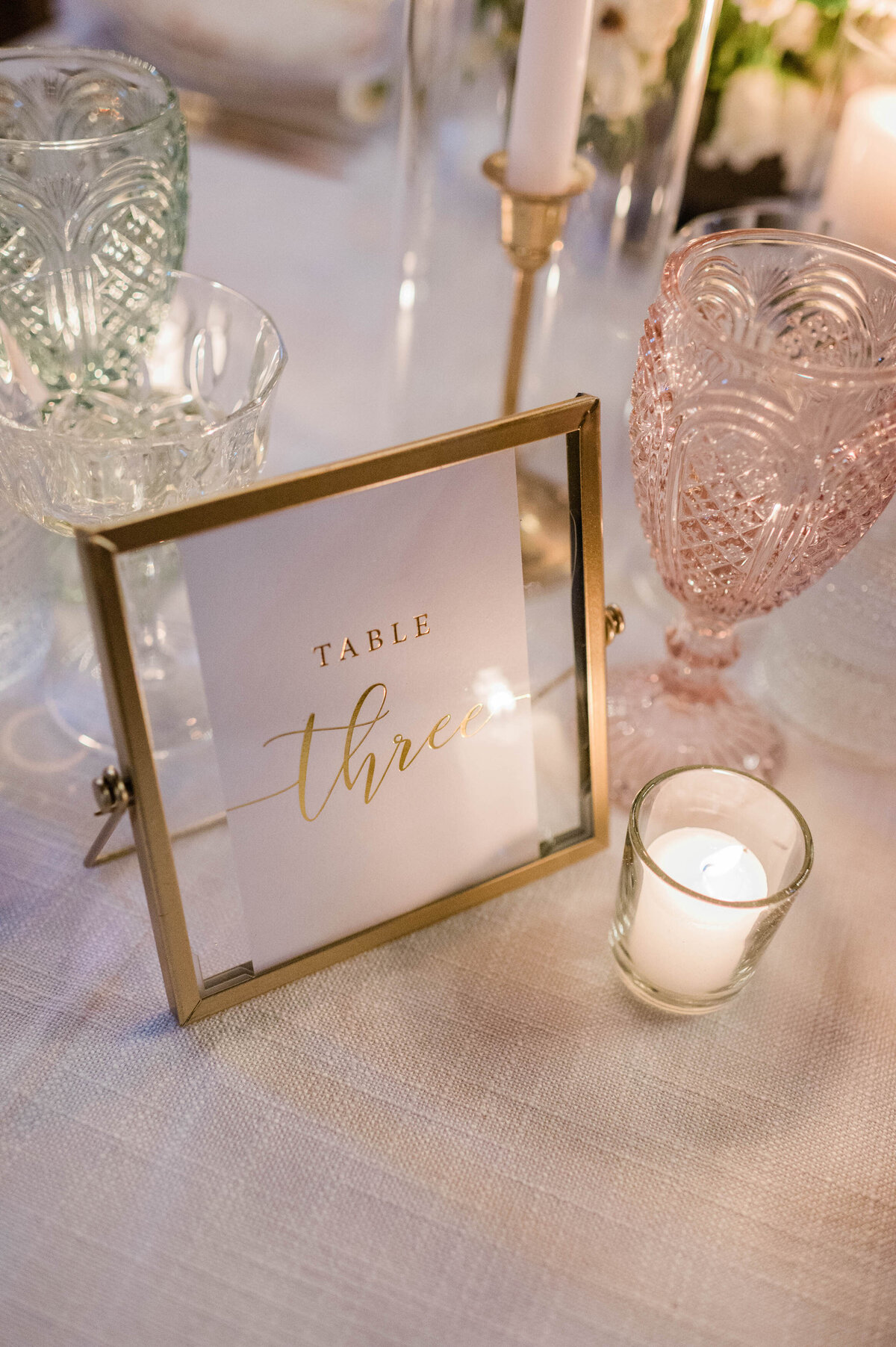 A gold-framed table number "three" surrounded by candlelight, vintage glasses, and fine tableware.