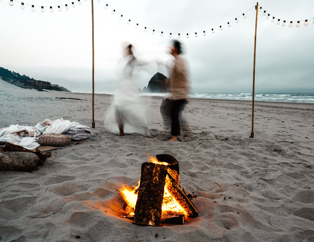 A blurry photo captures a couple dancing in front of a beach bonfire with Haystack rock in the background for their Oregon Coast elopement.