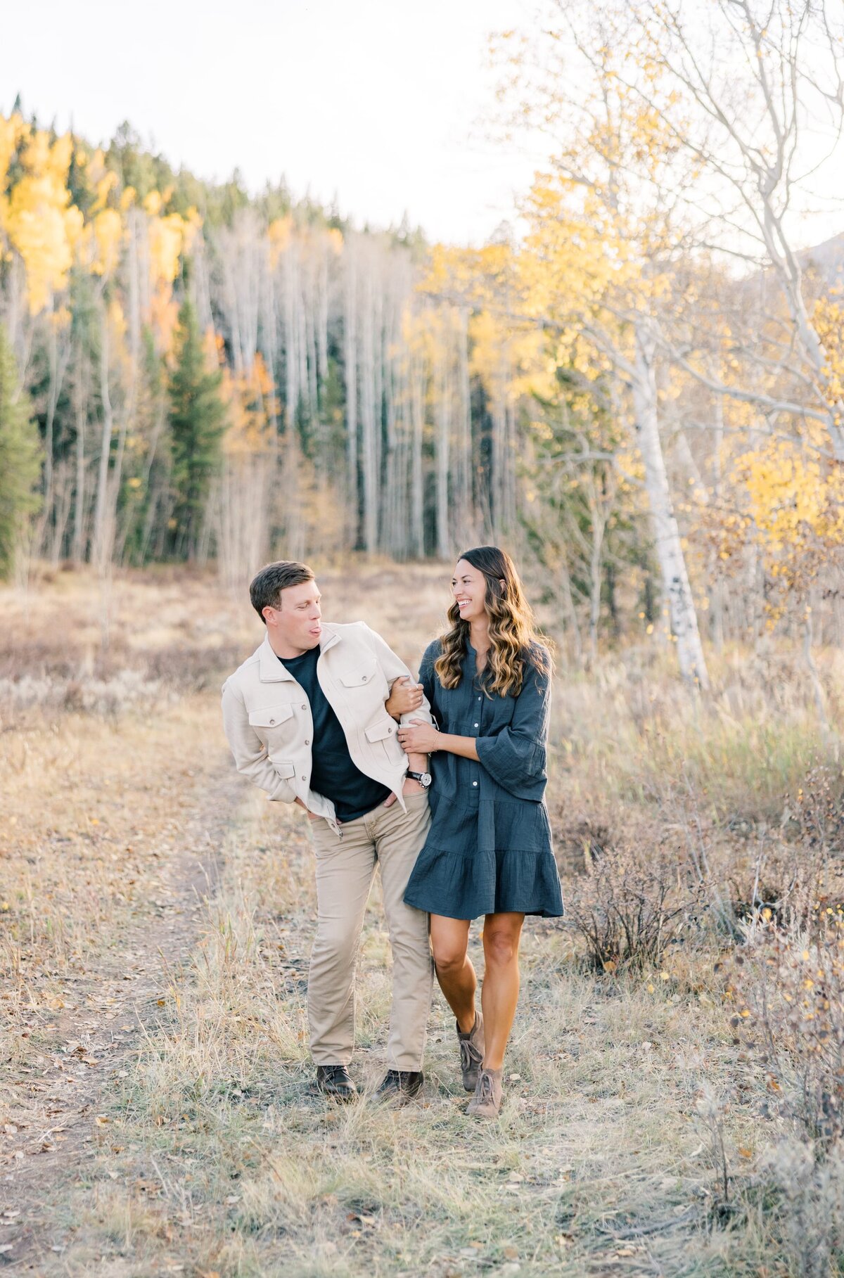That same couple is walking playfully together. They are bumping hips and laughing together. They are surrounded by yellow aspen trees in the fall. 