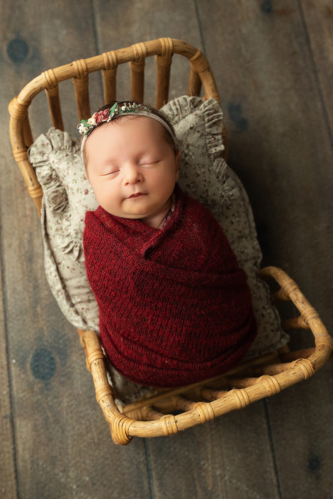 sleeping newborn swaddled in a deep red swaddle with a dainty floral headband as she is in a tiny baby sized bed for newborn photos 