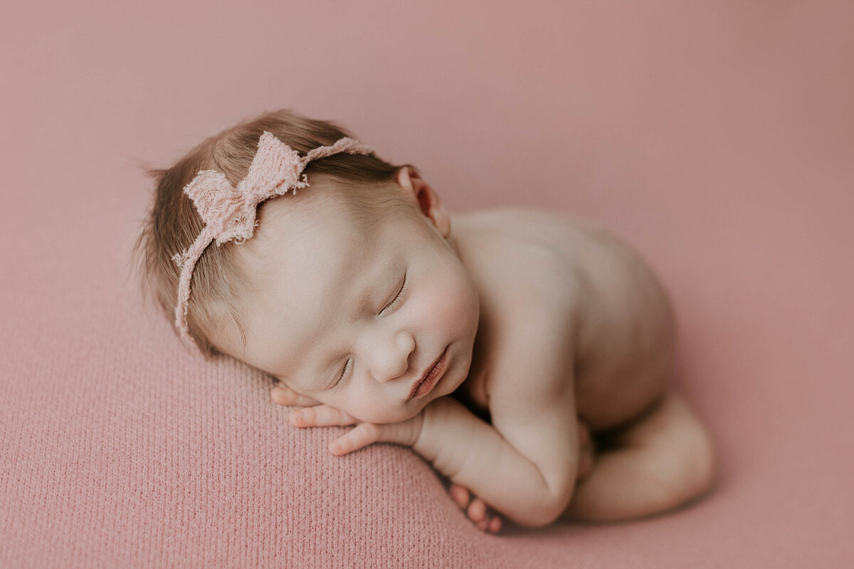 Studio newborn photography - Baby sleeping bare on top of a pink blanket. Baby is resting on top of their legs with their hand under their cheek.