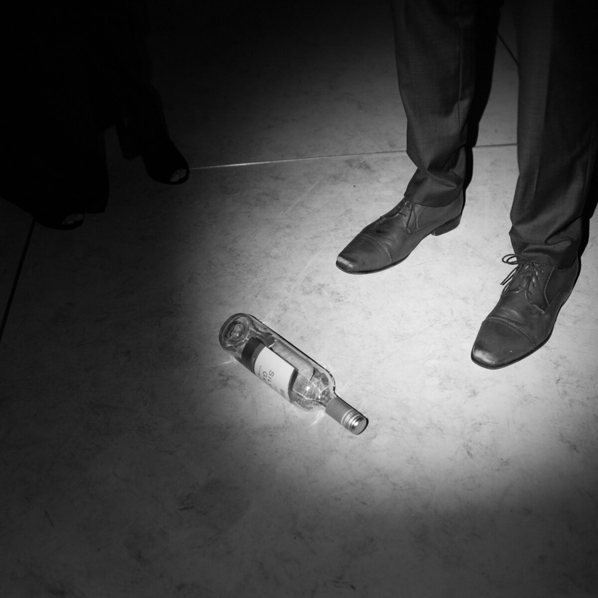 A black and white film detail of an empty bottle left on the wedding dance floor, capturing the raw, late-night energy of the celebration by Kate Winters