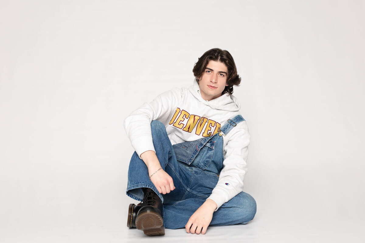 A person with long hair sits cross-legged on the floor wearing a white Denver hoodie and denim overalls. The background is plain white.