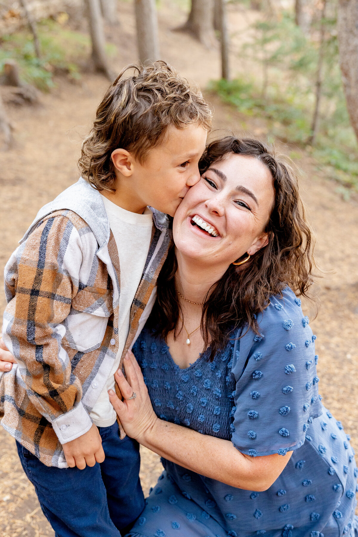 A mother in a blue dress kneels as her young son leans in to give her a kiss on the cheek, both laughing joyfully in a forest setting.
