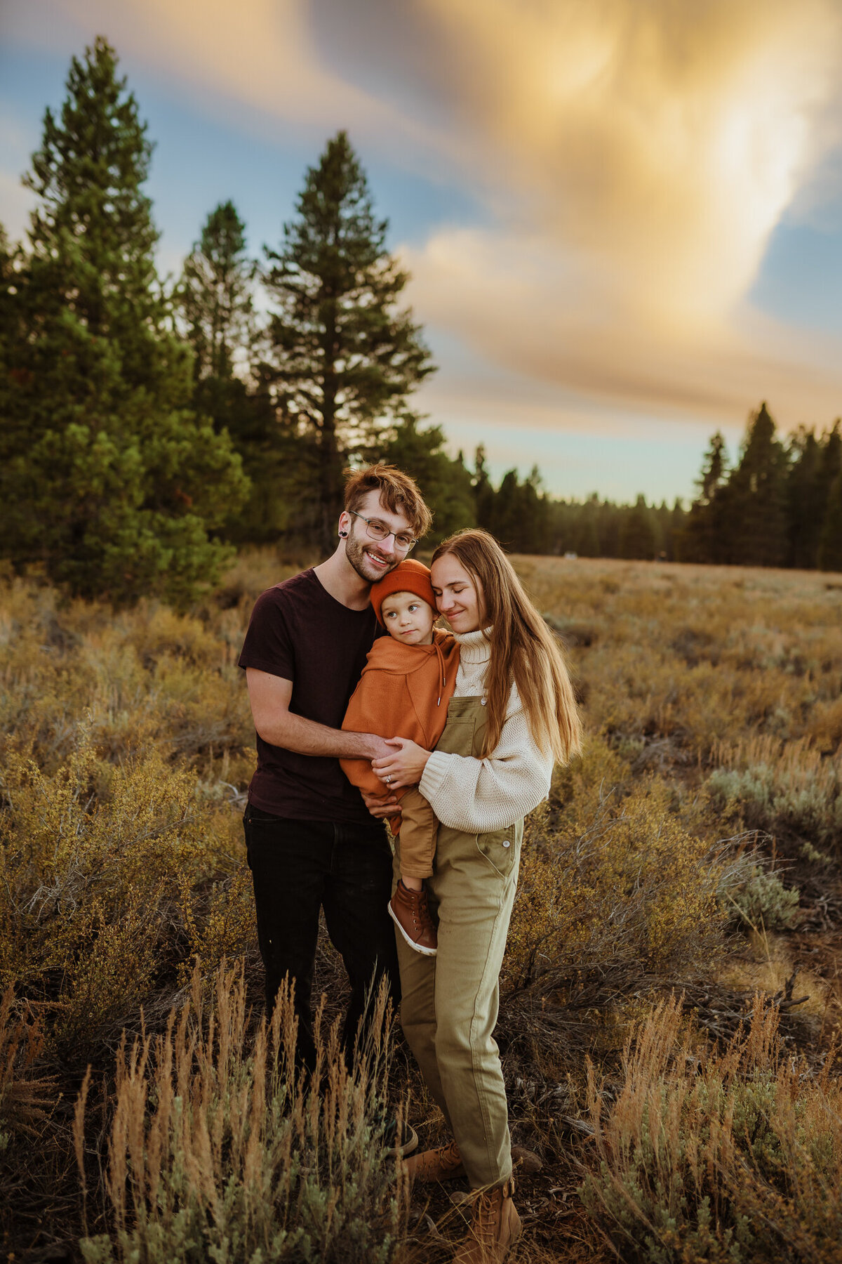 Mom and dad are standing and holding their toddler son and snuggling him with a beautiful sunset and trees behind the,