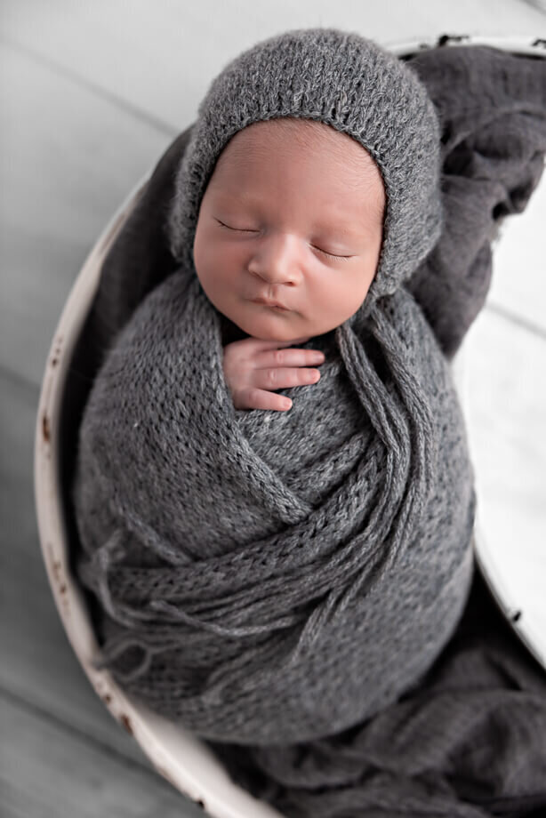 newborn baby boy in a denim blue wrap with a matching bonnet.  He is posed on his back in a white moon bowl