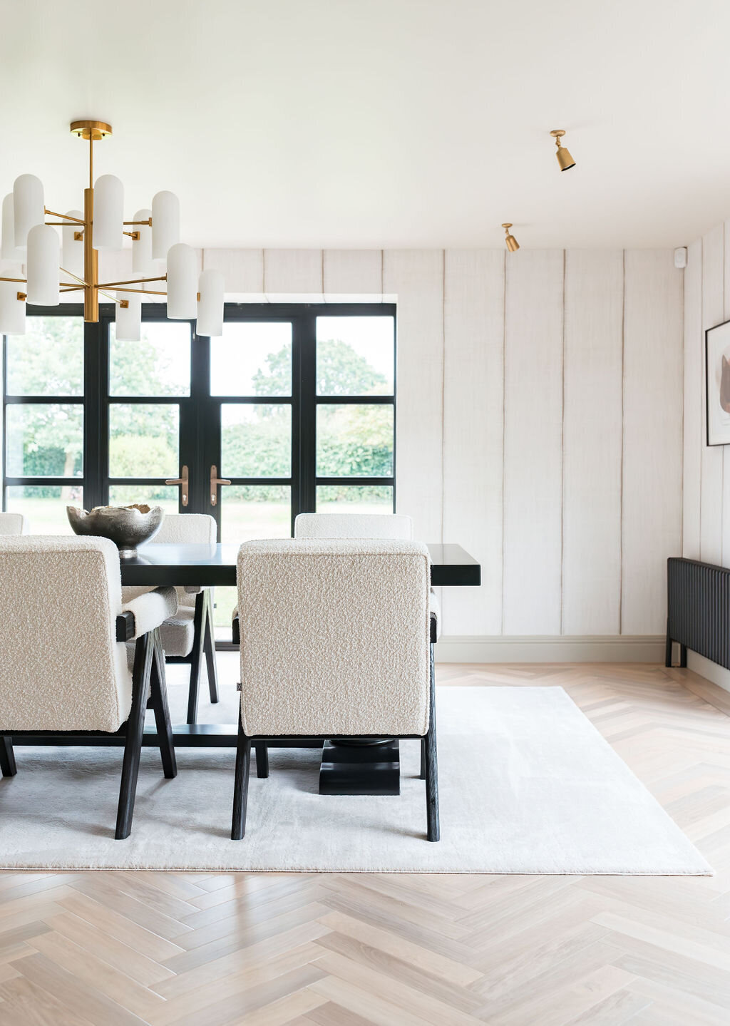 A modern dining room features a sleek black table surrounded by white upholstered chairs with black legs. A large white area rug covers the herringbone-patterned wooden floor. There is a gold chandelier and abundant natural light flooding through large windows.