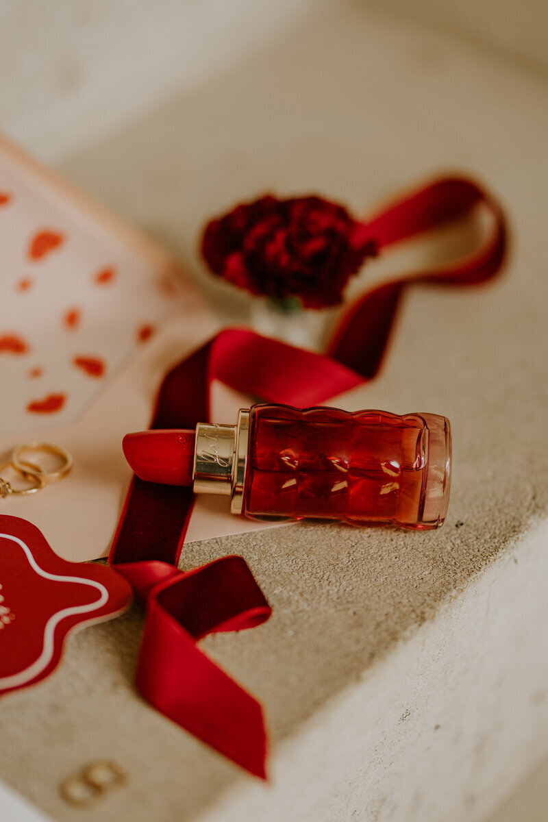 Mise en scène imaginée par Laura, photographe mariage en Vendée, dans laquelle sont délicatement positionnés un rouge à lèvre, un ruban rouge, des alliances et de la papeterie.