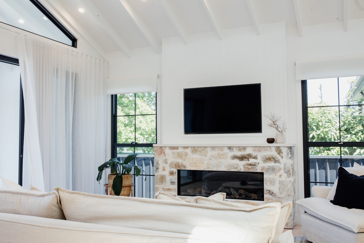 Modern living room in Newcastle with white walls, vaulted ceiling, stone fireplace, TV, and large windows. Cozy beige sofas enhance the bespoke interior design that adds warmth and sophistication to the space.