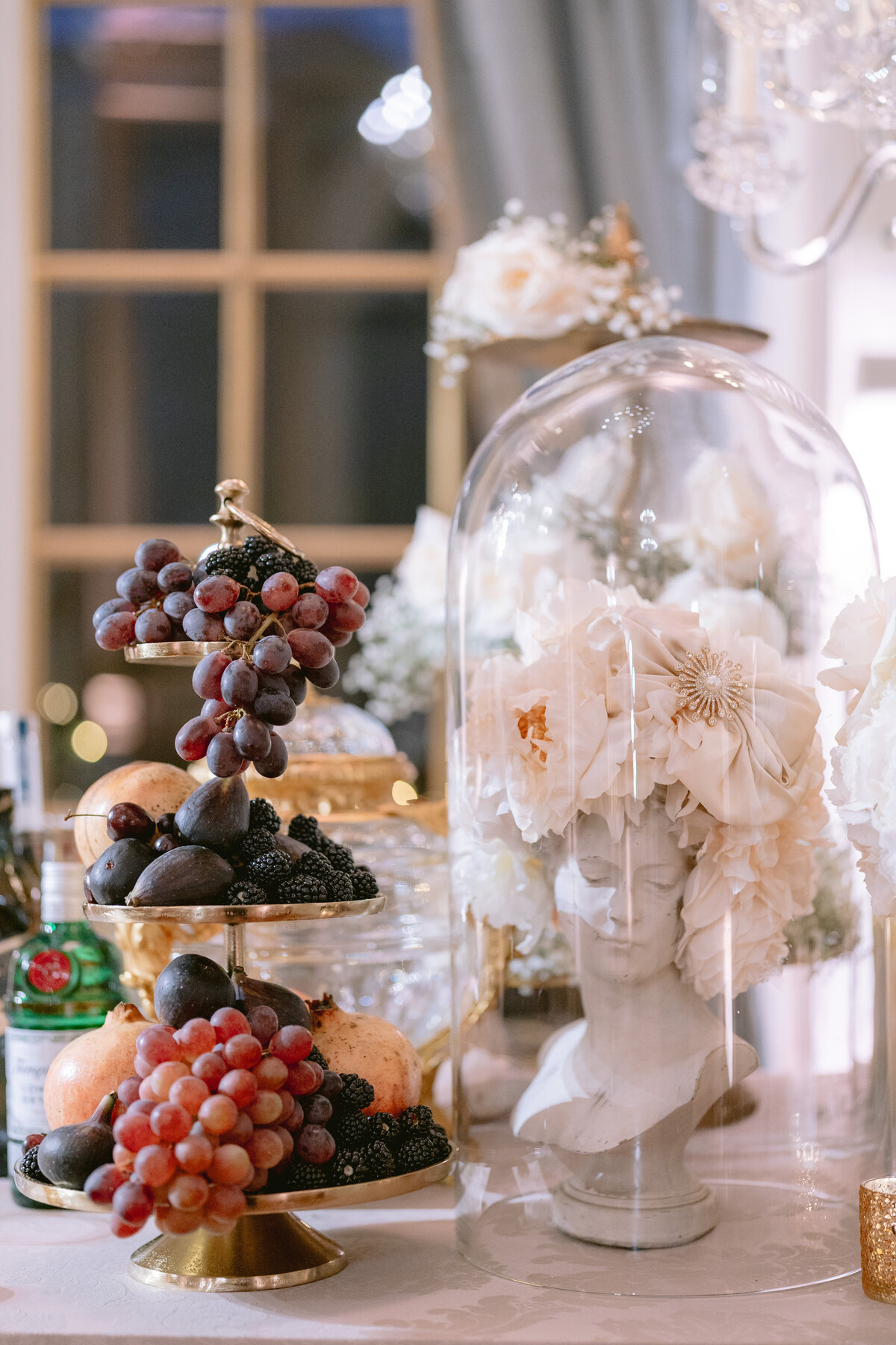 The photo was taken at the luxurious Shangri La Hotel & Ritz Paris Hotel in Paris, France, capturing a lavish display meticulously arranged by photographer Matteo Coltro and videographers Zen Film Works. The centerpiece of the display is a tiered tray showcasing an opulent array of red and black grapes, luscious figs, and juicy pomegranates. Adjacent to this is a striking white statue bust embellished with delicate white flowers, elegantly encased in a glass dome for added charm. The aesthetic is further enhanced by the presence of additional floral accents, a picturesque window backdrop, and sophisticated decor elements, creating a truly captivating visual experience.