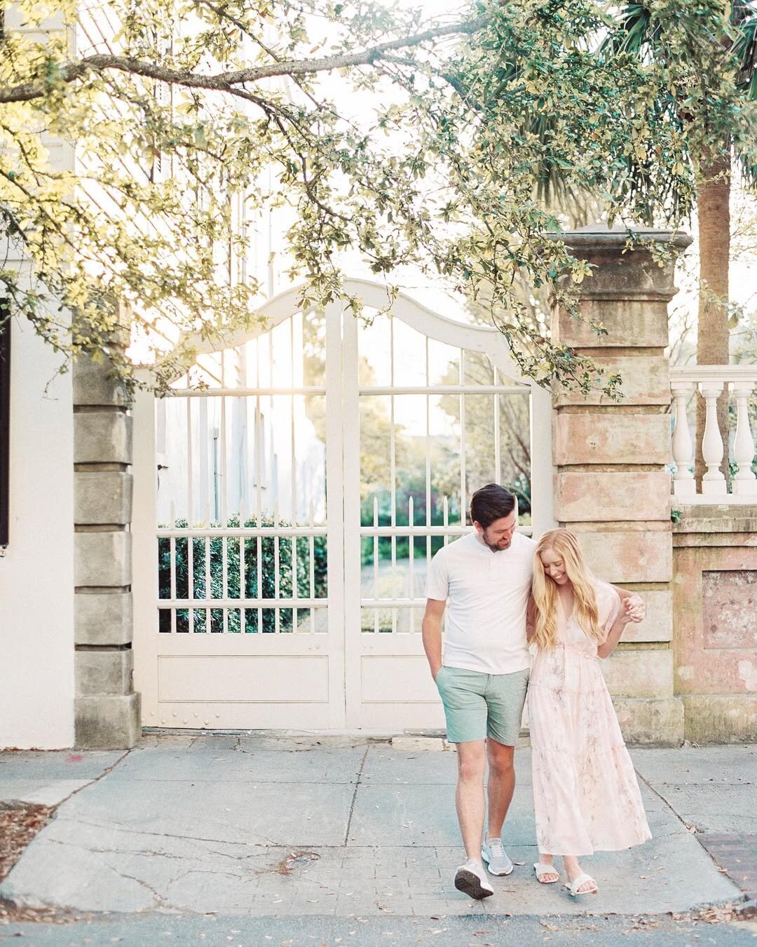 Wedding Photographer RI | A couple strolls arm in arm, smiling, in front of a white gate. The setting is sunlit, with leafy trees overhead.