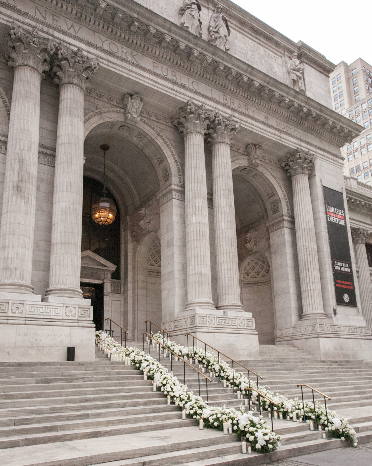 The stunning photo was captured at the grand entrance of the New York Public Library in New York City by the talented photographer Christian Oth. The scene features the iconic columns and steps of the library, elegantly adorned with beautiful white floral arrangements. The historic building boasts intricate carvings, while the steps are lined with handrails that lead the way up to the imposing large arched doors. Additionally, the videography for this event was beautifully captured by East West Films.