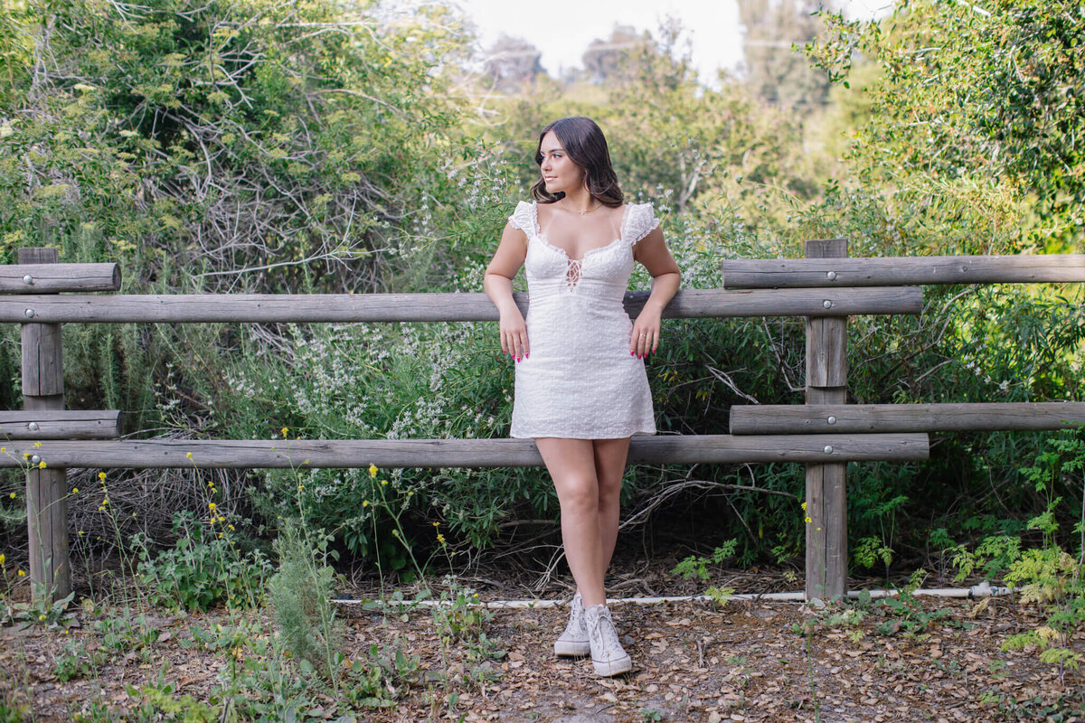 A woman in a white dress stands by a wooden fence in a lush, green outdoor setting. She is looking to the side, with one hand on the fence and the other resting on her hip. She is wearing white sneakers.