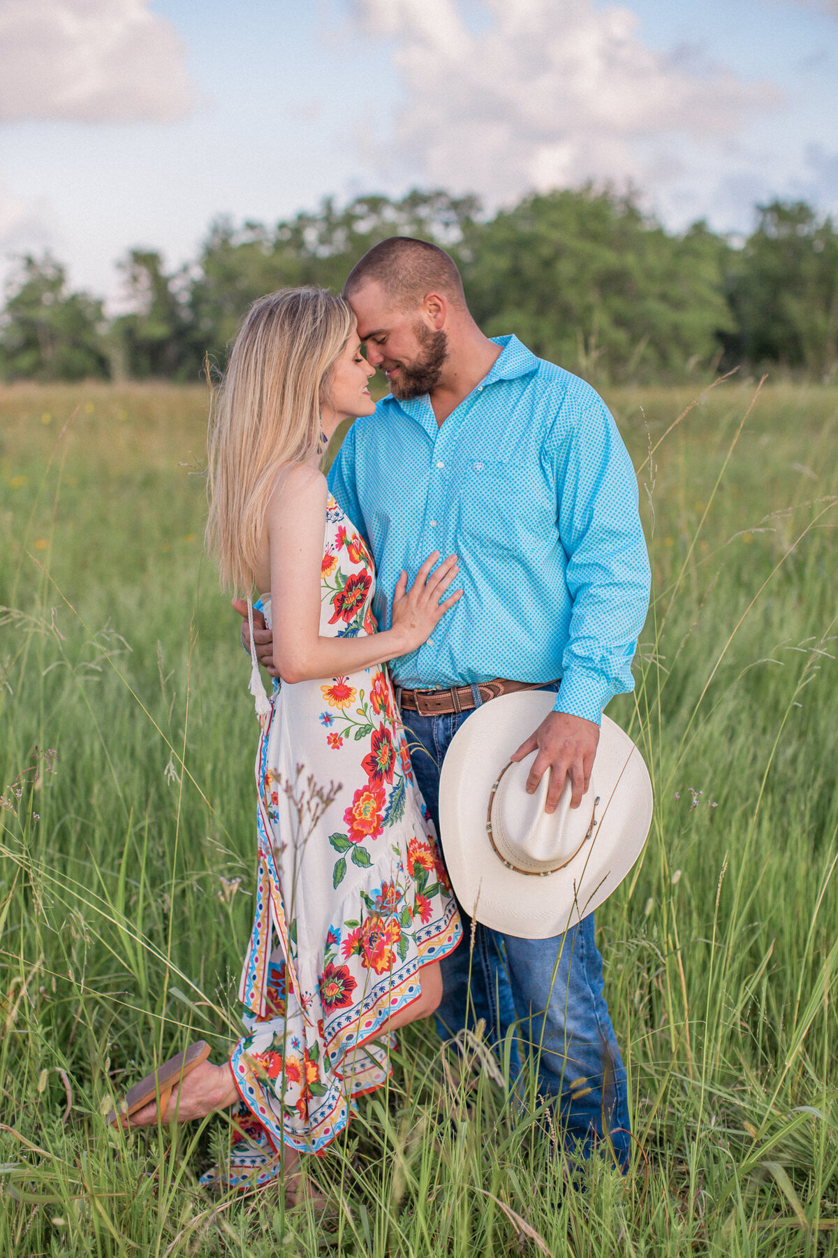 A blond woman in a floral dress snuggles close to her fiance, who is holding a white cowboy hat