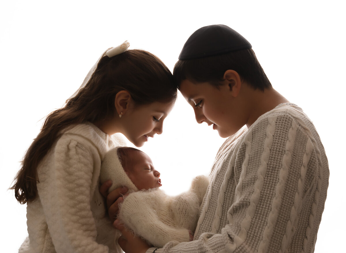 Brother and Sister hold their new baby brother. Brother and sister are facing each other and big brother is holding baby. brother and sister's heads are touching and they are looking down at baby. Captured by top Brooklyn newborn photographer Chaya Bornstein