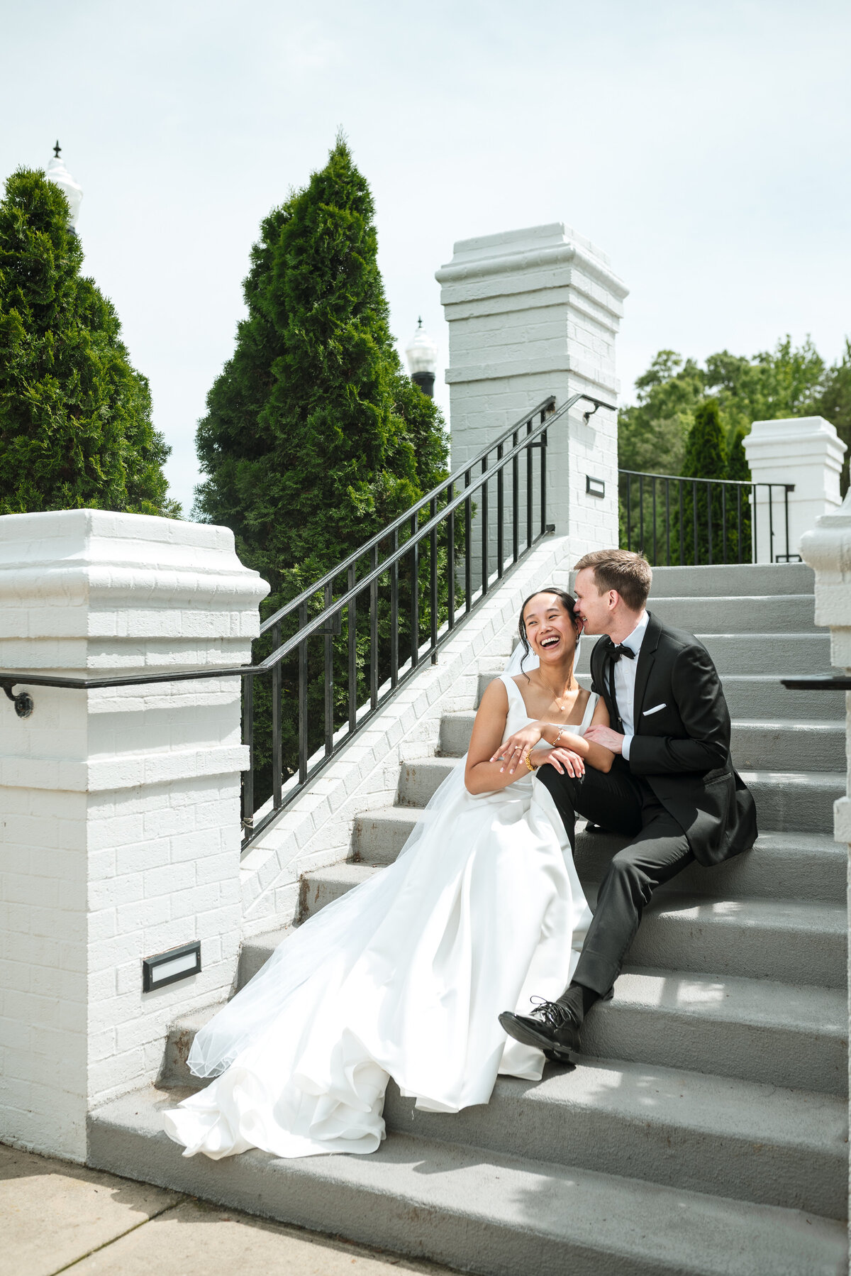 Romantic wedding photo at Providence Country Club in Charlotte, NC featuring a joyful bride in a timeless white gown and groom in a classic black tuxedo sharing a candid, laughter-filled moment on the staircase. Captured by Charlotte wedding photographers DeLong Photography, specializing in luxury weddings and country club venues.