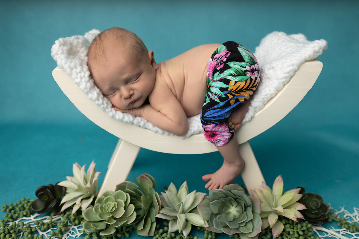 Close-up of sleeping baby boy wearing vibrant floral pants posed on a white bench with greenery.