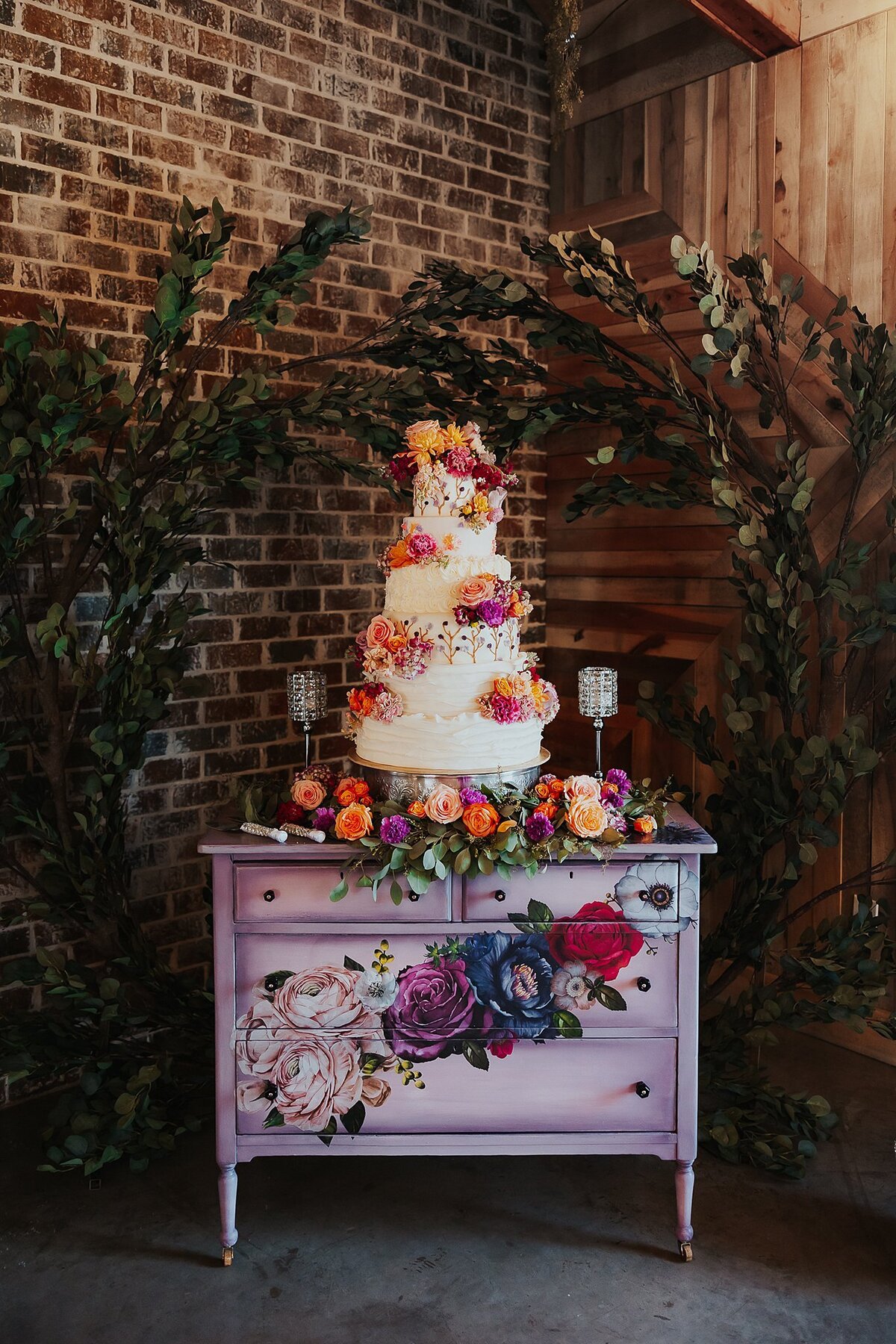 An extra large seven tier wedding cake decorated with purple roses, orange roses, peach roses, and blush roses sits on a purple dresser with lilac roses, purple roses and red roses painted on the front. Behind the cake is an exposed brick wall and two arching ficus trees creating an arbor. ,