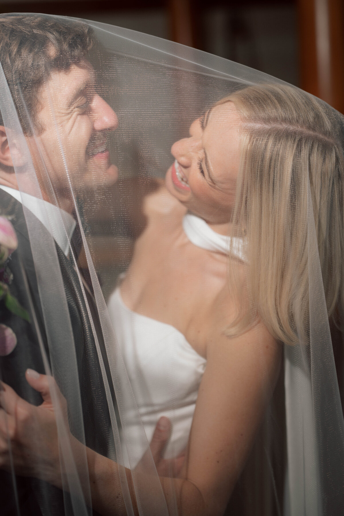 Bride and groom laughing together under veil in intimate wedding portrait