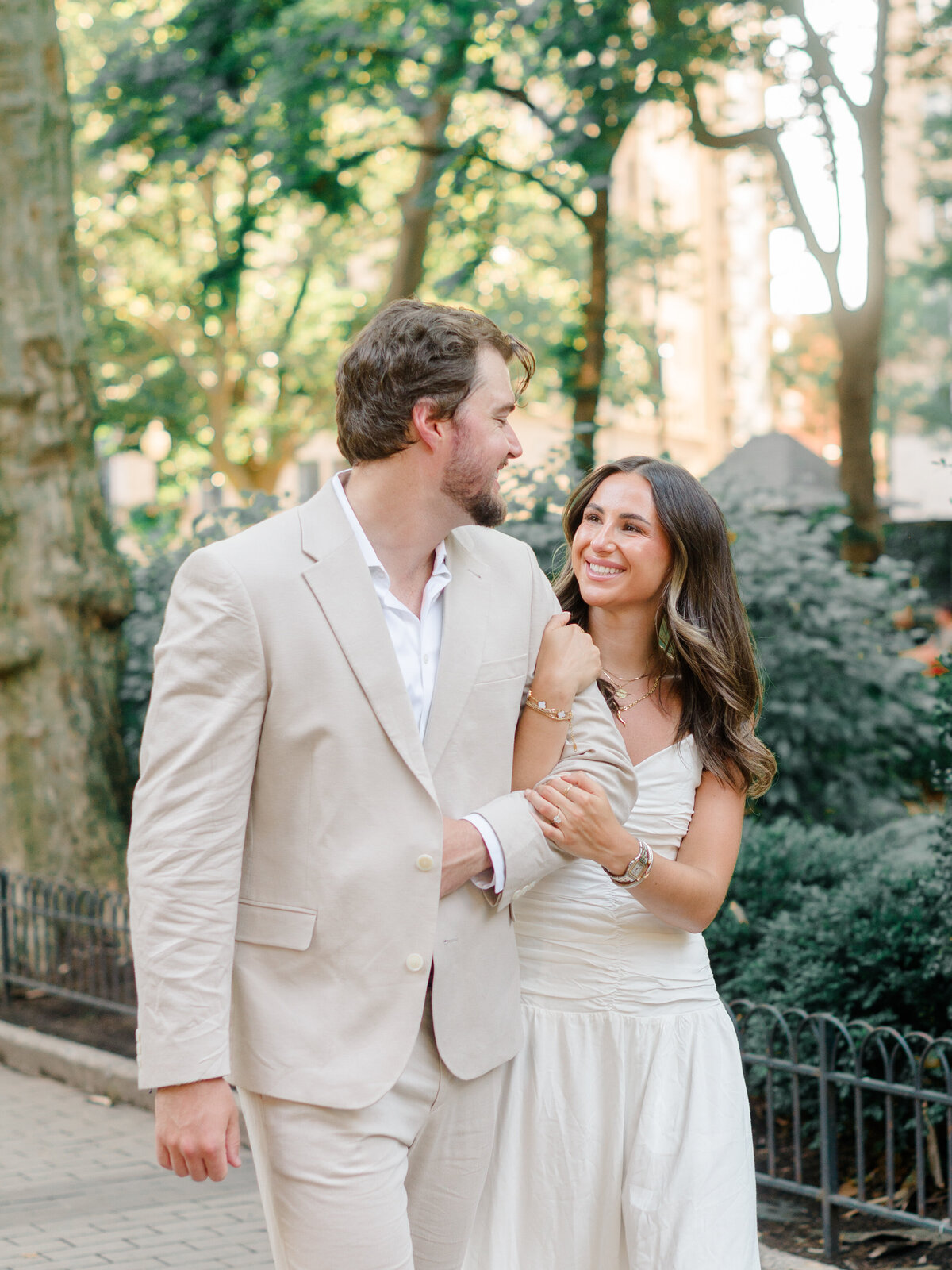 A couple strolls arm in arm through Rittenhouse Square, sharing a lighthearted moment under the trees. Dressed in soft neutrals, they smile at each other with warmth and ease, perfectly capturing the charm of a Philadelphia engagement. Photographed by Elizabeth Kane Photography, a Luxury Philadelphia Wedding Photographer.