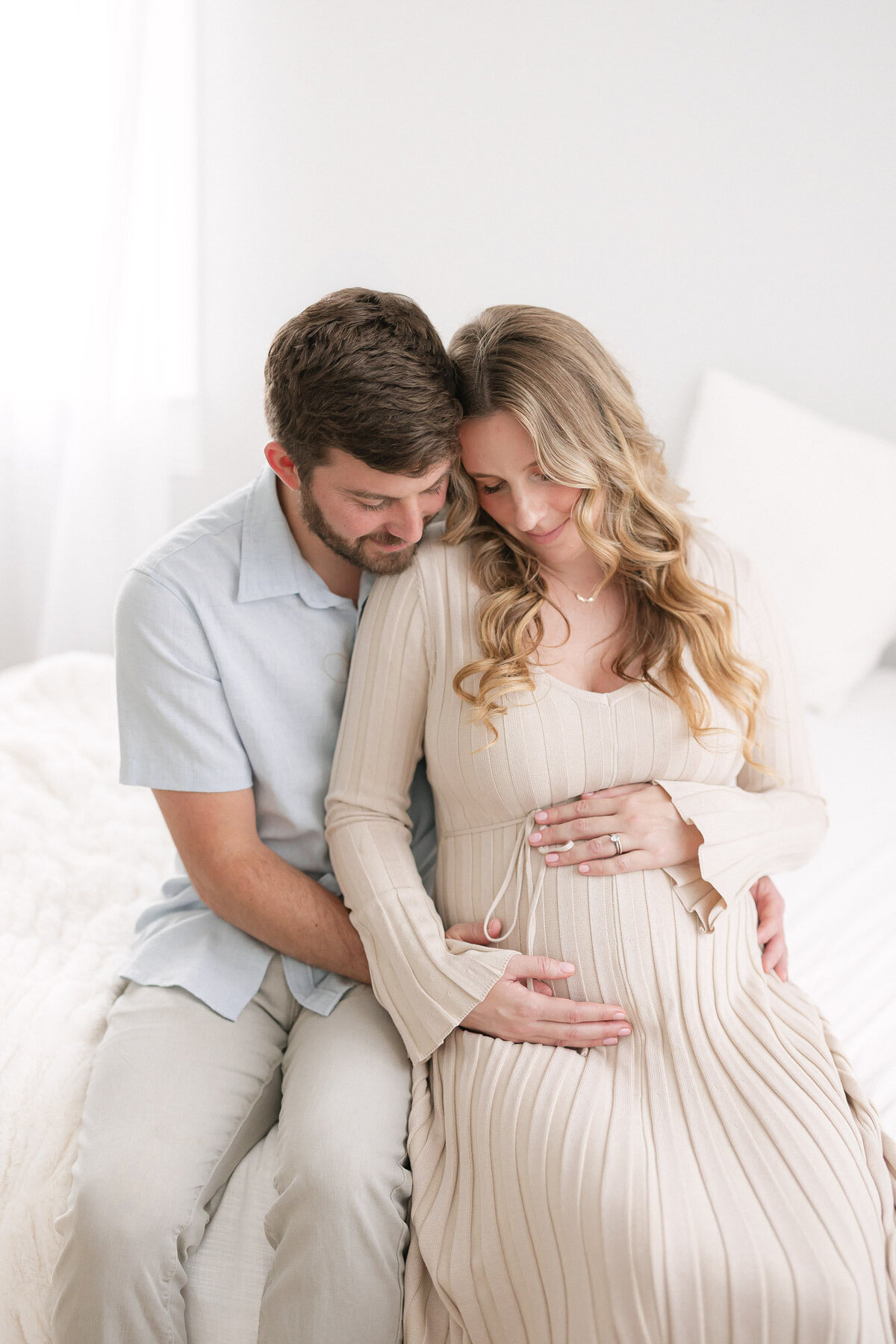Portrait of a pregnant mom and her husband sitting on a white bed as they embrace her baby bump in missy marshall photography's bright louisville kentucky studio